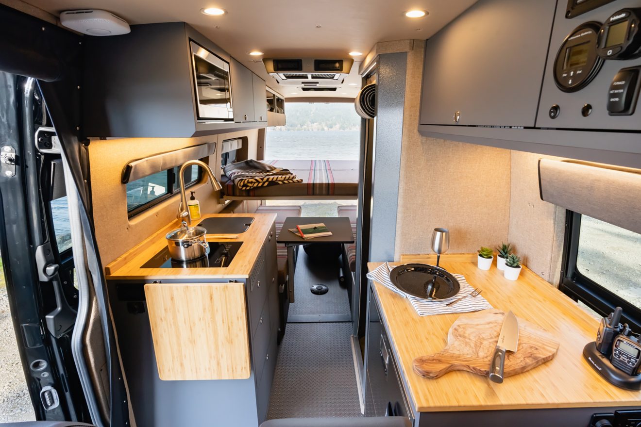 Sprinter van interior, large galley kitchen with flip-up table on the left, fully enclosed aluminum shower on the right, next to a stainless steel refrigerator