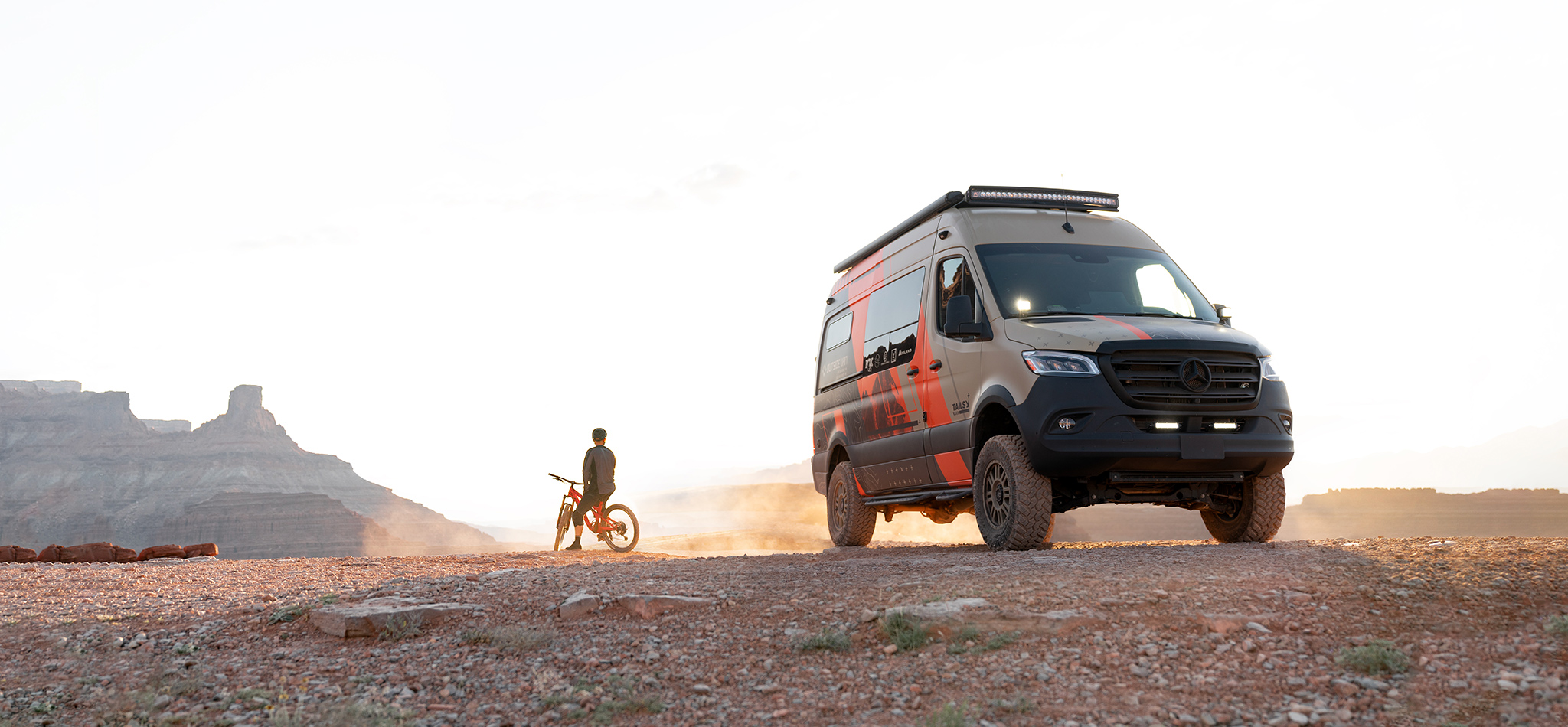 A mountain biker looks out over the desert beside a custom Sprinter camper van.
