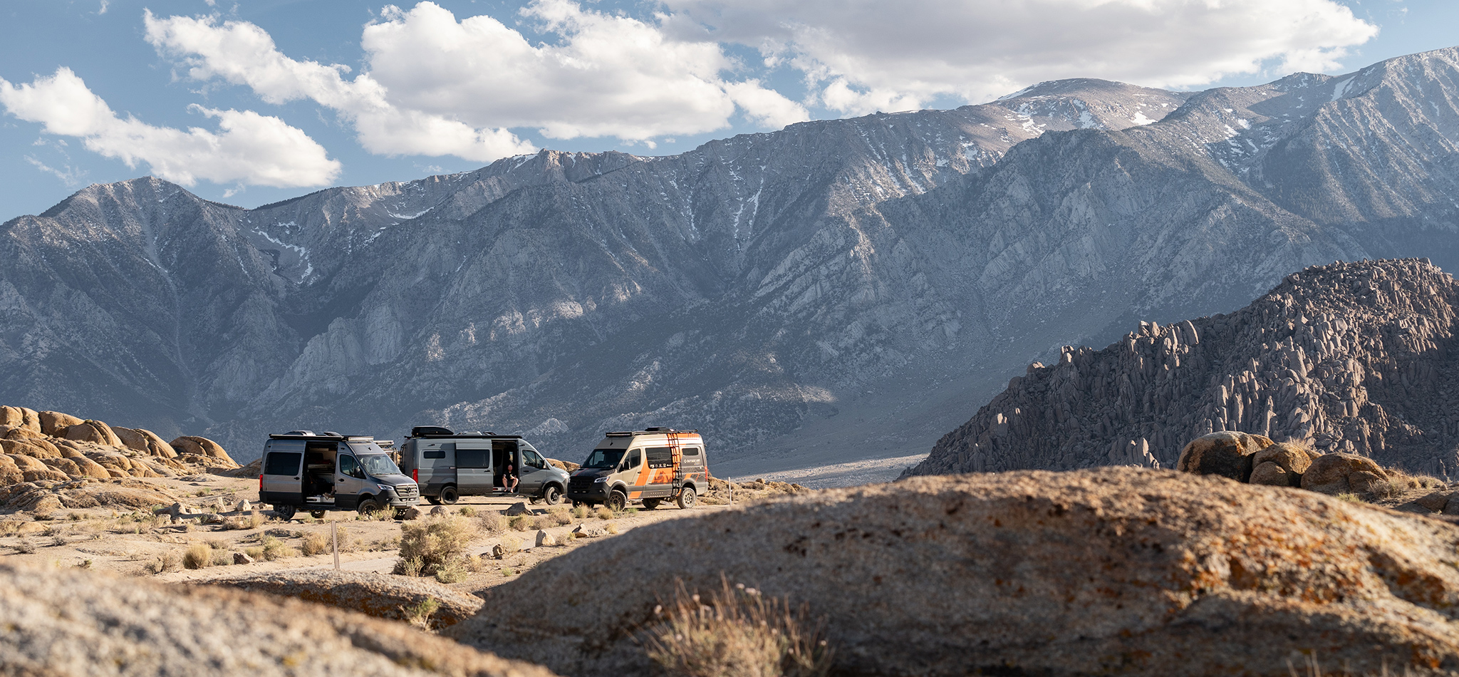 Three custom Sprinter camper vans are circled up at a campsite with desert mountains in the distance.