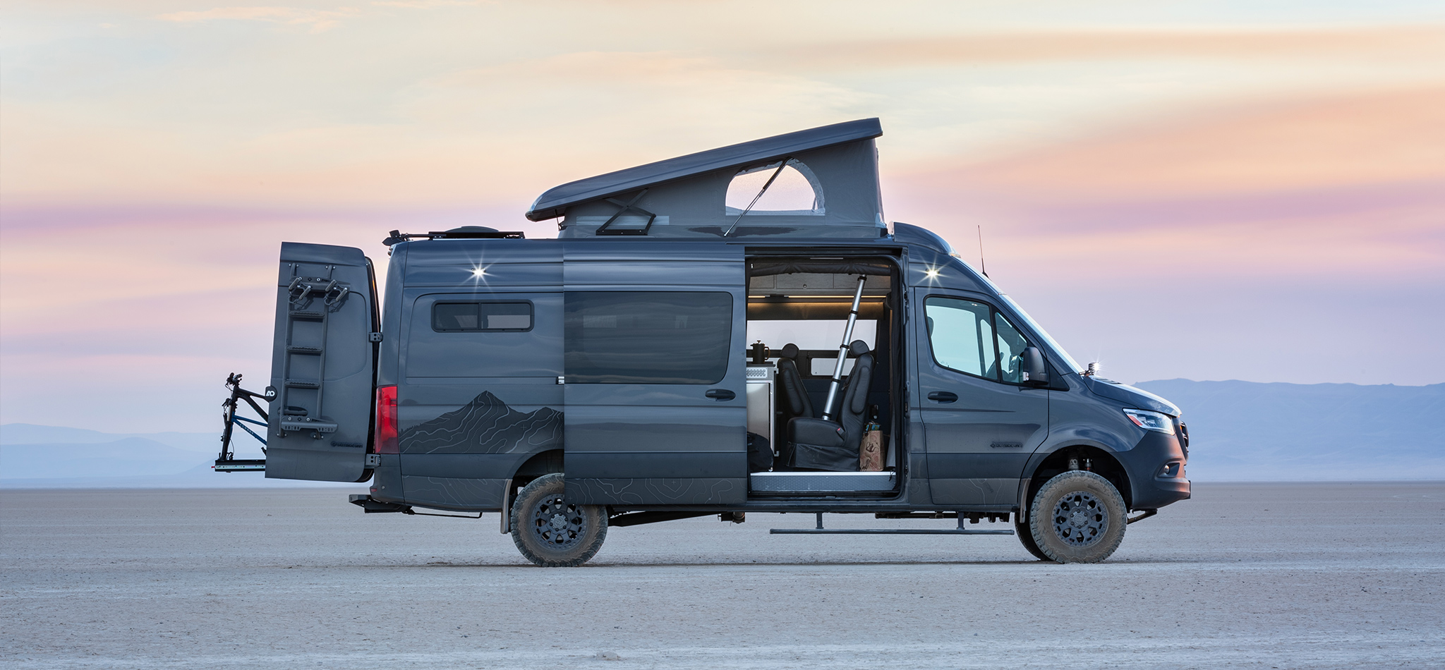 A Sprinter van conversion with a roof top tent and bicycle racks rests on top of a playa at dusk.