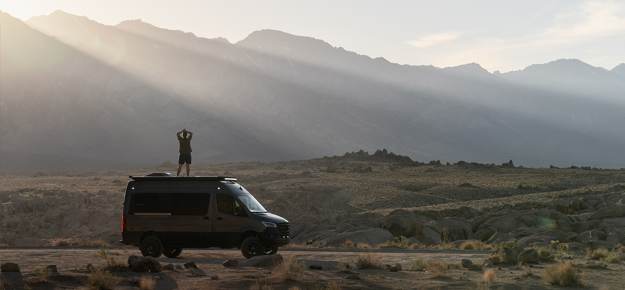 A person stands on top of their custom Sprinter van looking at the sunlight breaking over mountain tops in the distance.