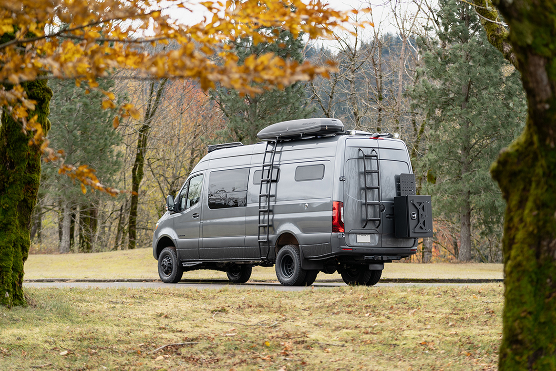 Exterior shot of a grey van with a rooftop box, driver-side ladder, rear door ladder, and storage box in a green forest