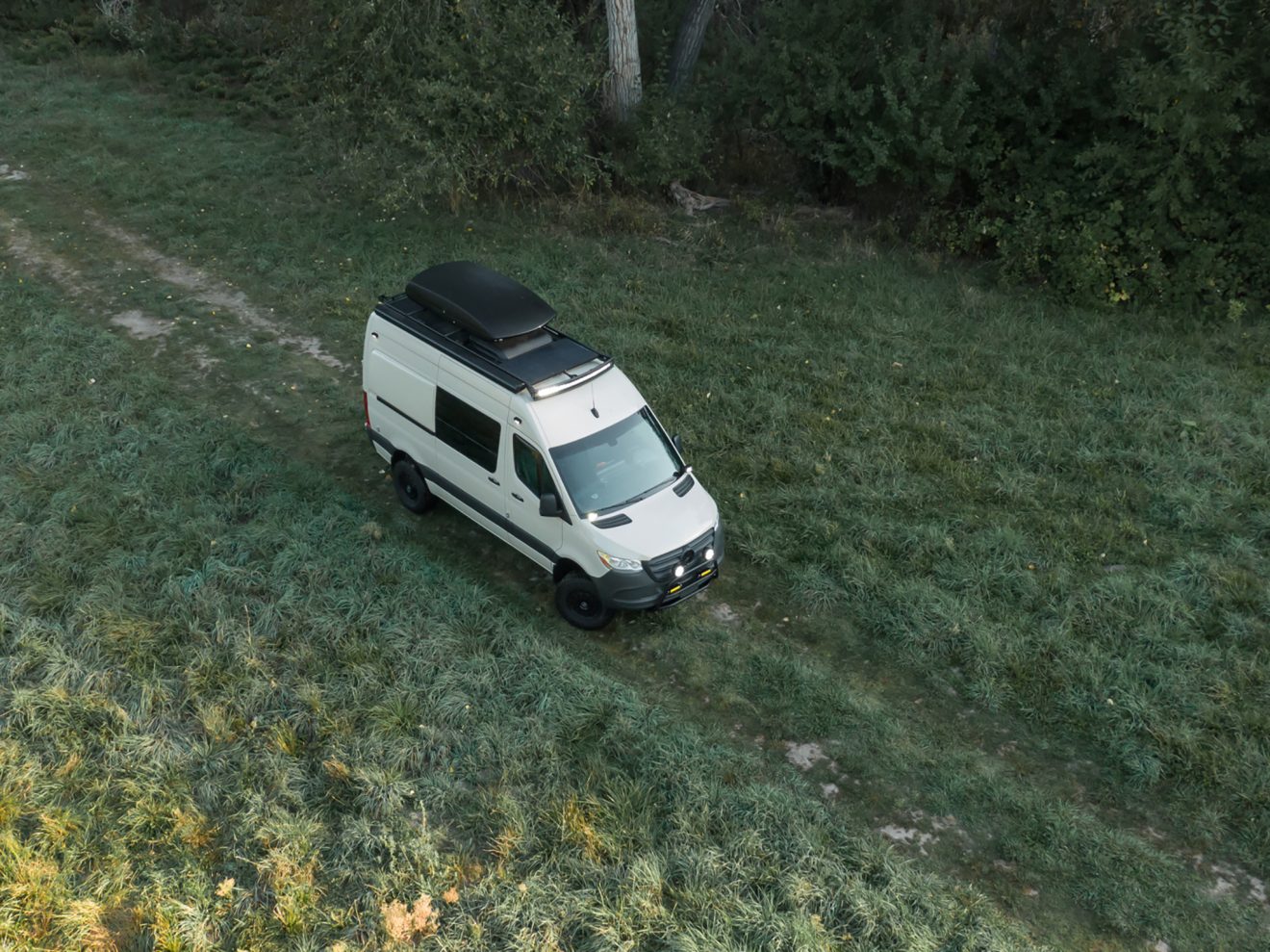 Aerial drone photo Exterior of a 2021 Mercedes-Benz Sprinter 144 with rooftop box and side ladder in a green field