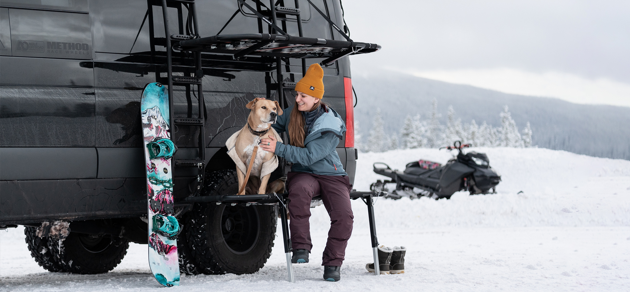 A woman smiles and pets her dog sitting on the edge of her customized Sprinter van conversion, with a snowboard and snowmobile nearby.