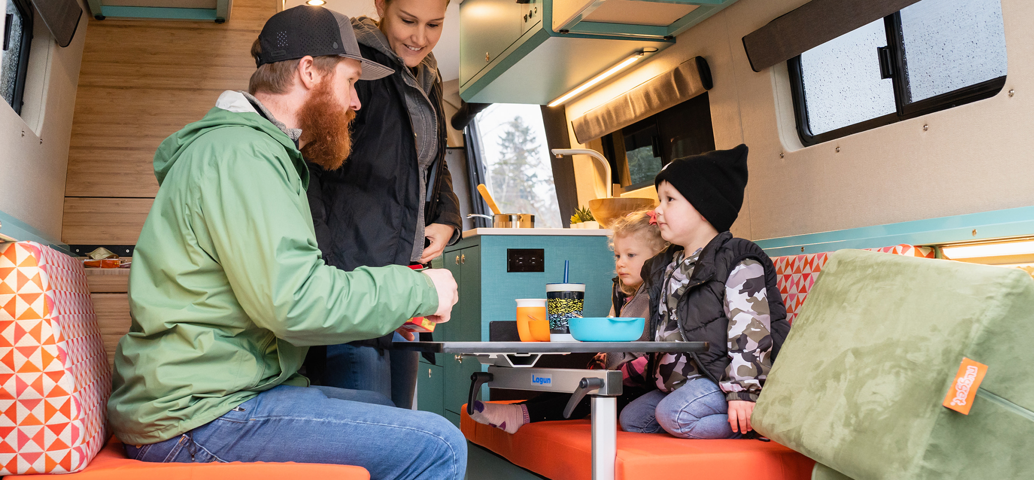 A family sits at a table and prepares to eat a meal in the comfort of their custom Sprinter camper van. It's raining outside.