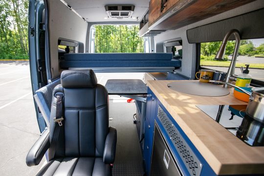 Wide Interior Image of custom off road sprinter van with table, kitchen, and blue and dark wood cabinetry
