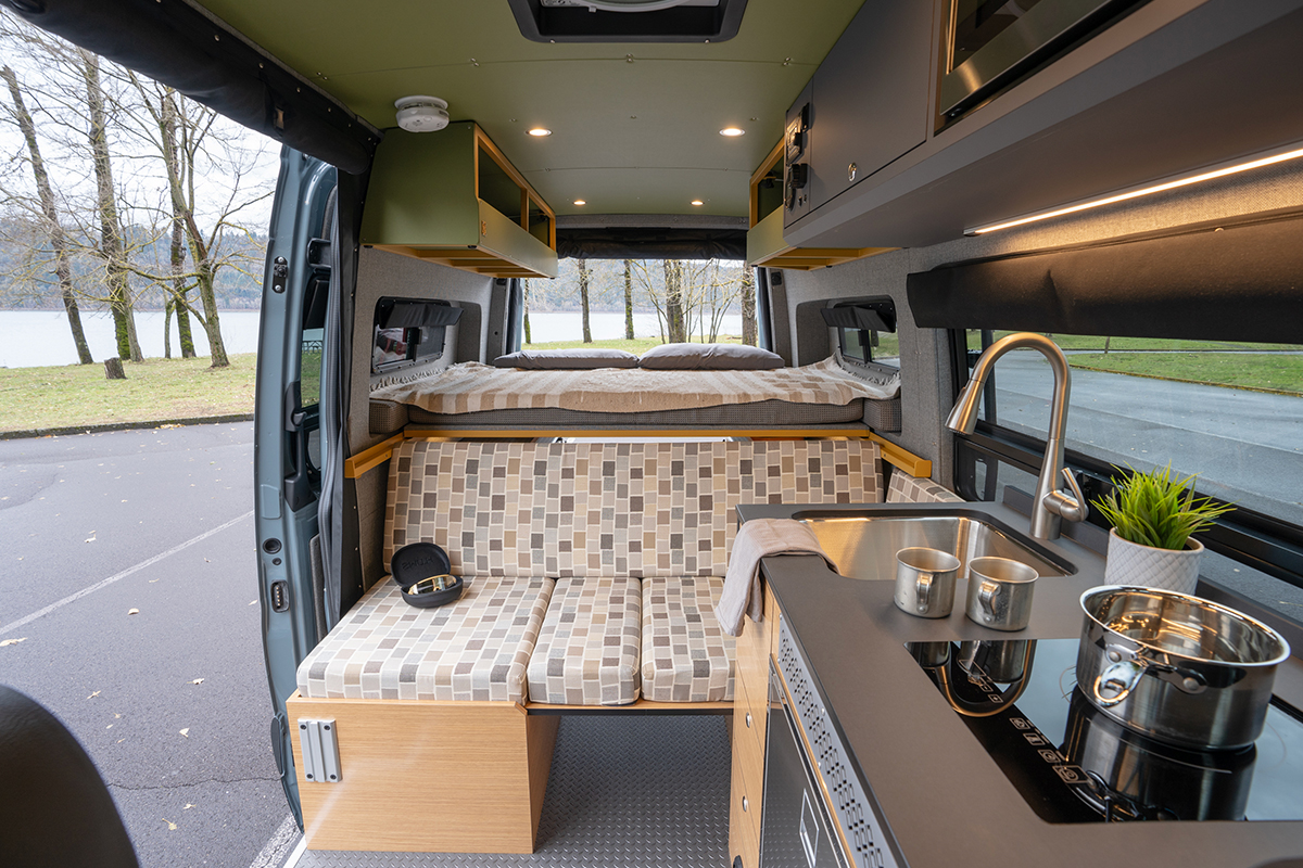 Custom-built van interior with bench sofa, passenger slider door open, kitchen with sink and burner on the right