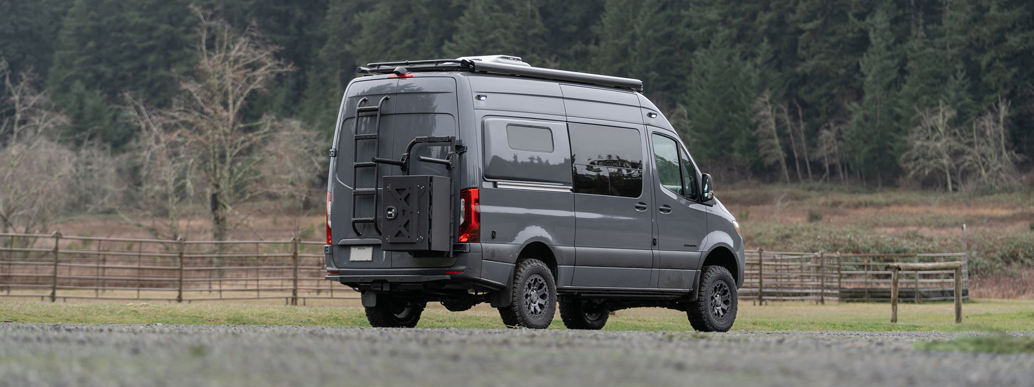 Grey van in a field with exterior metalwork on rear panels with a ladder, cargo box, and bike rack