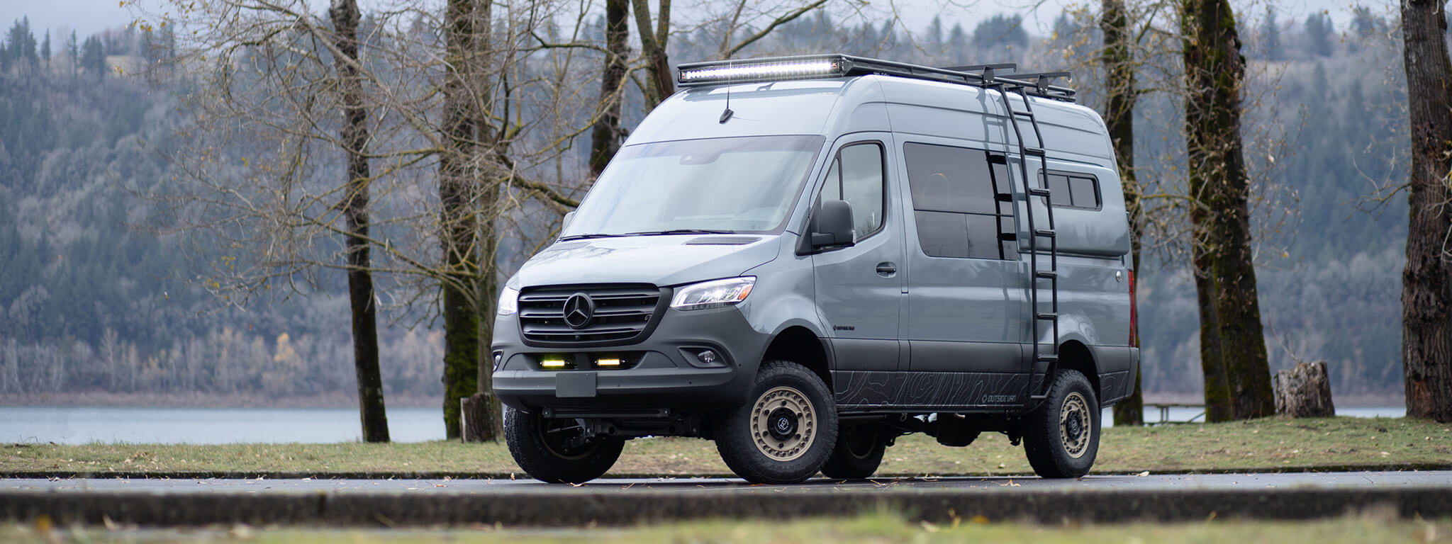 Exterior blue mercedes-benz van with driver-side utility ladder in parking lot with trees and lake