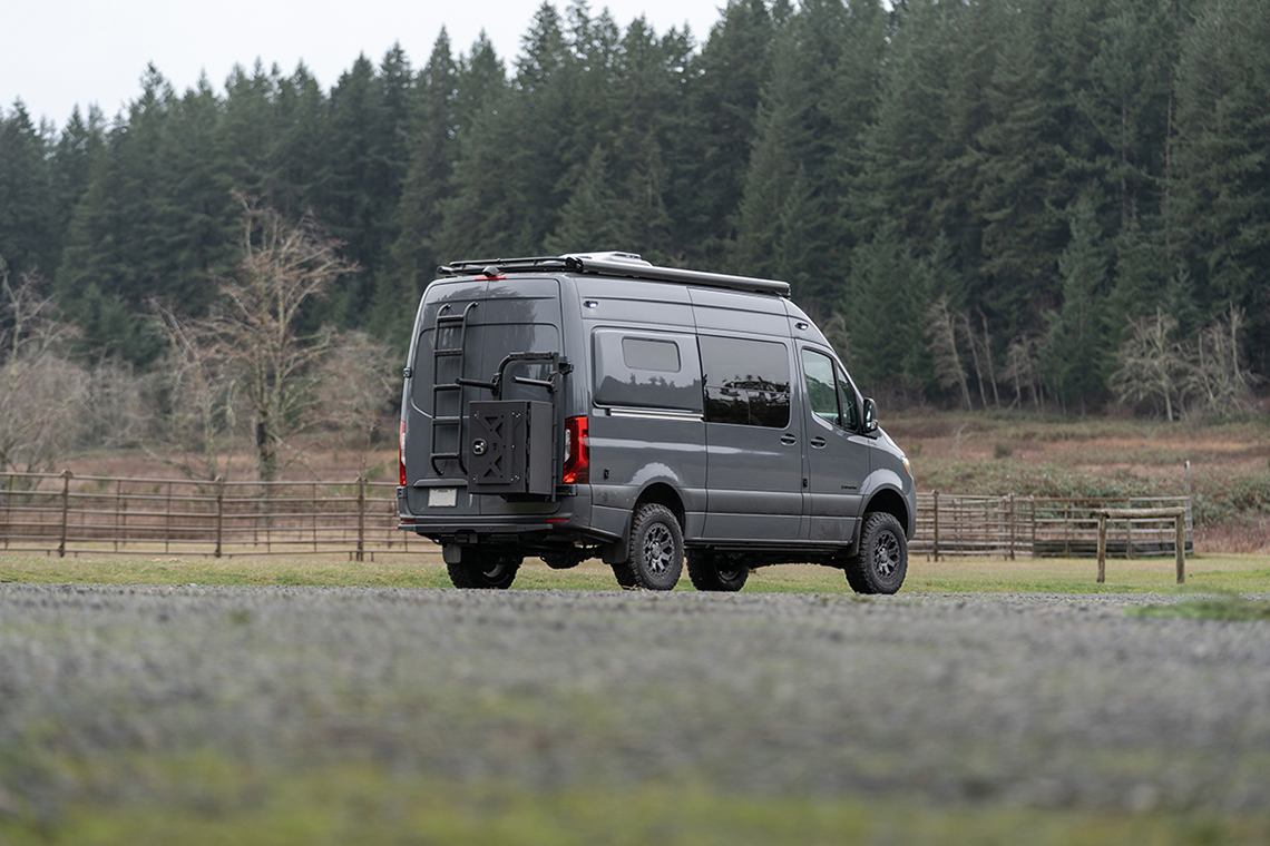 Grey van in a field with exterior metalwork on rear panels with a ladder, cargo box, and bike rack