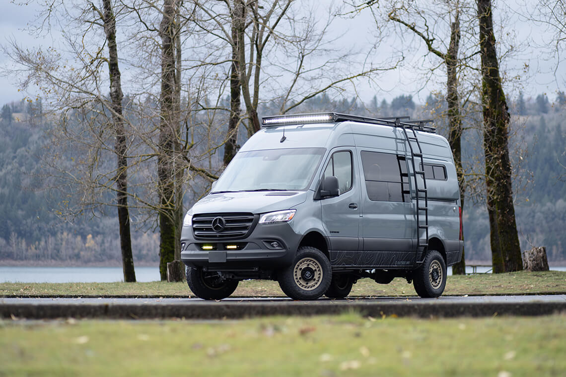 Exterior blue mercedes-benz van with driver-side utility ladder in parking lot with trees and lake
