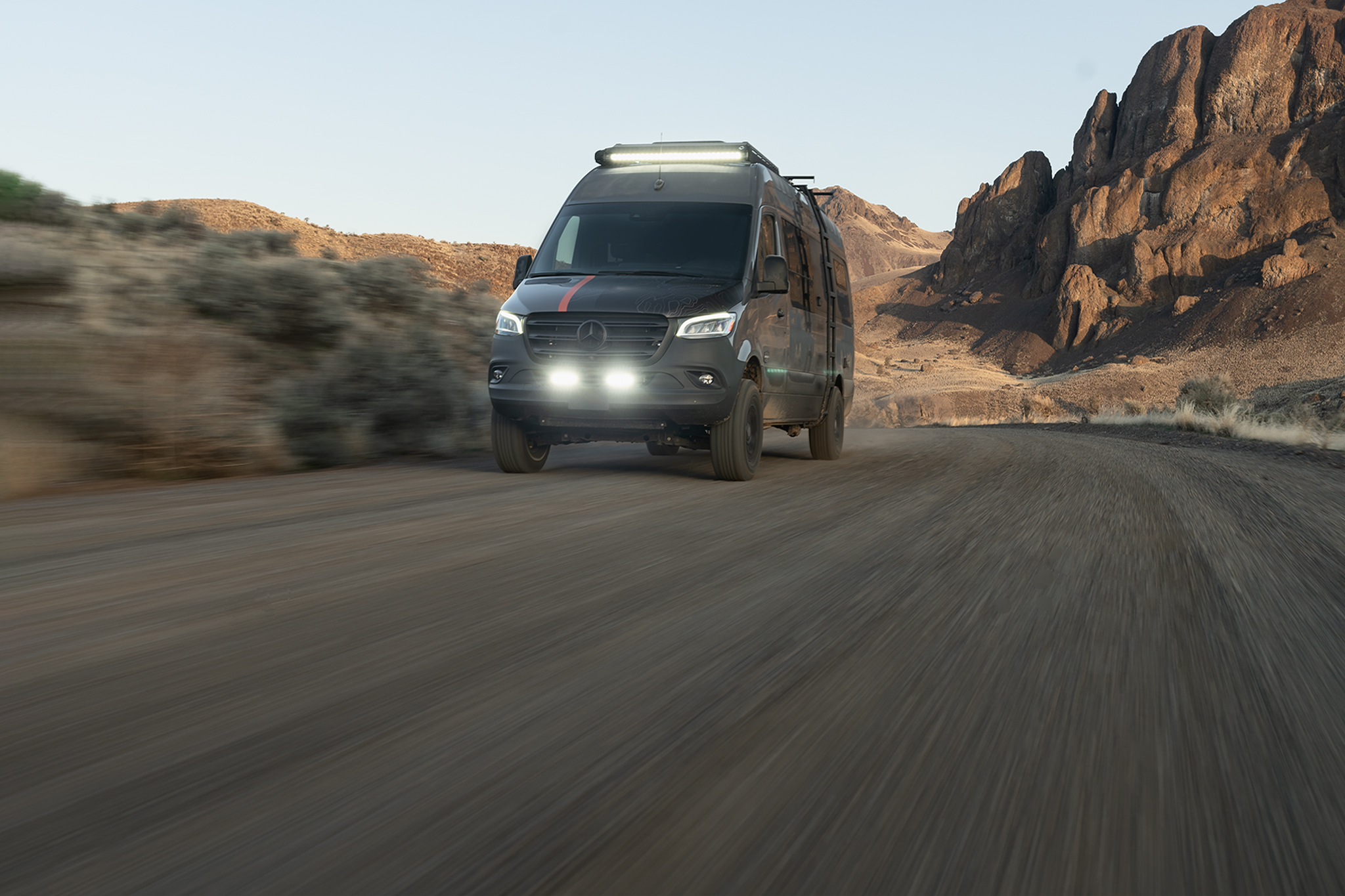 A 4x4 Sprinter adventure van drives along an off-road trail through a gulch. Its light bars illuminate the road.
