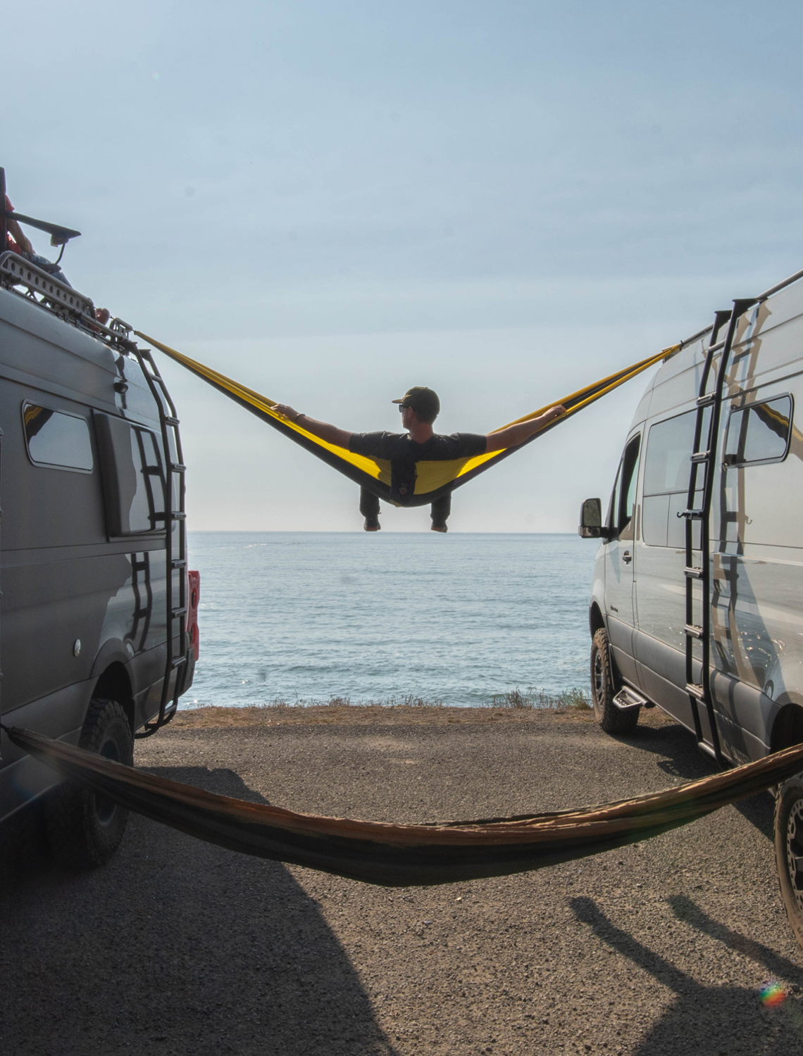 A man sitting in a hammock stretched between two vans looking out over the ocean.