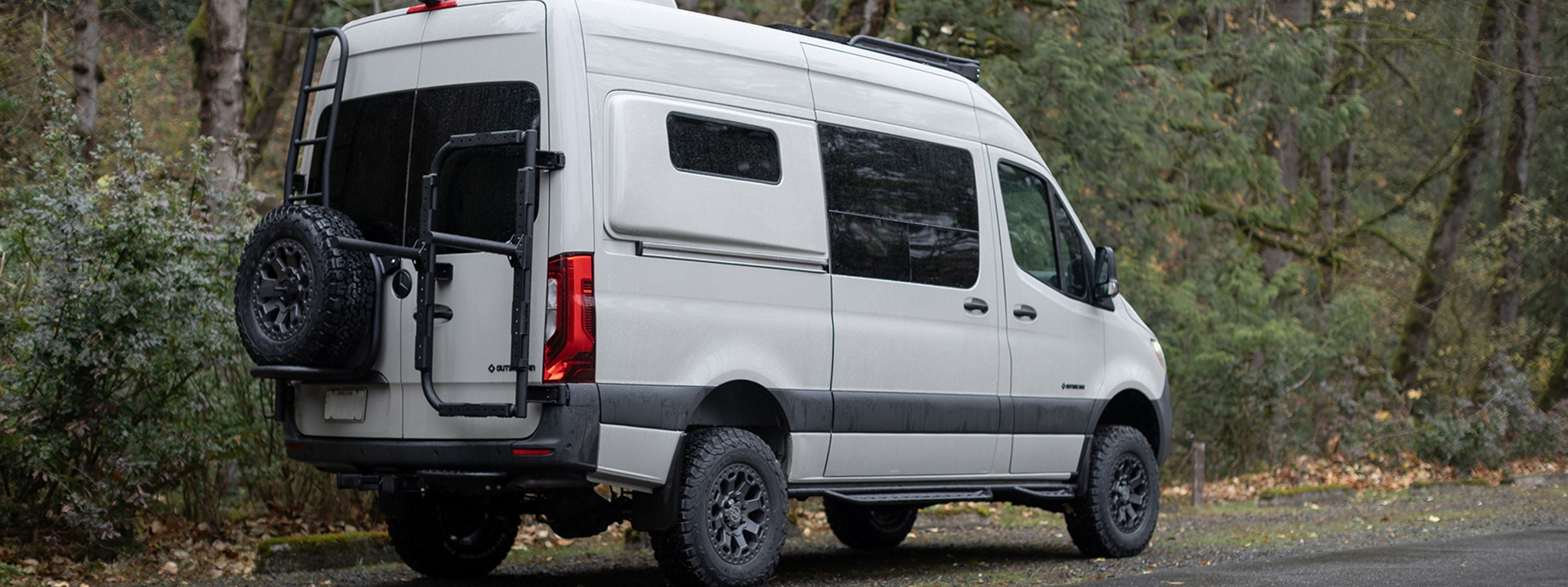 White van with rear ladder and spare tire carrier parked in a forest