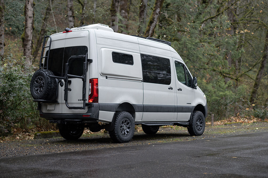 White van with rear ladder and spare tire carrier parked in a forest