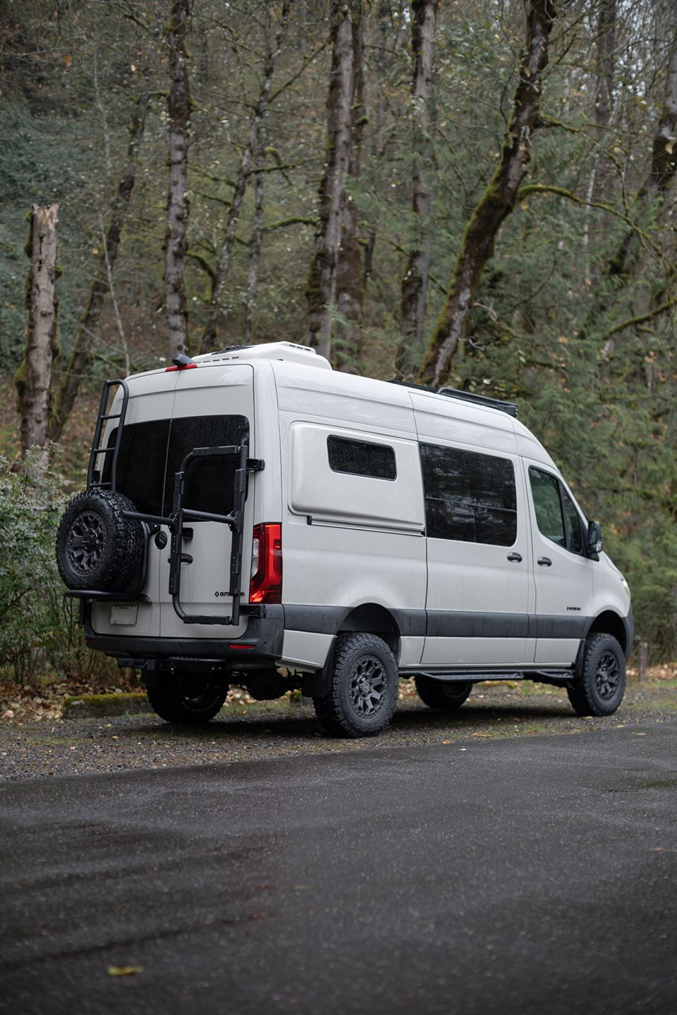 White van with rear ladder and spare tire carrier parked in a forest