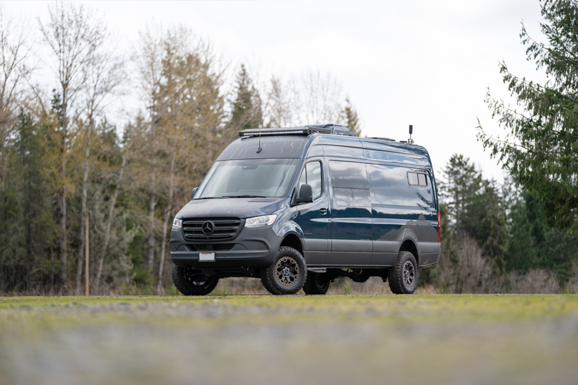 Custom off road mercedes sprinter van with blue exterior and topo wrap parked on gravel lot with trees in background