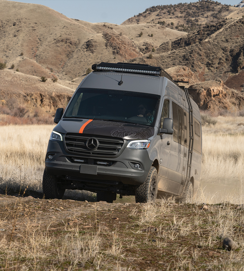 An off-road Sprinter conversion van drives over a small hill on a trail through some grasslands.